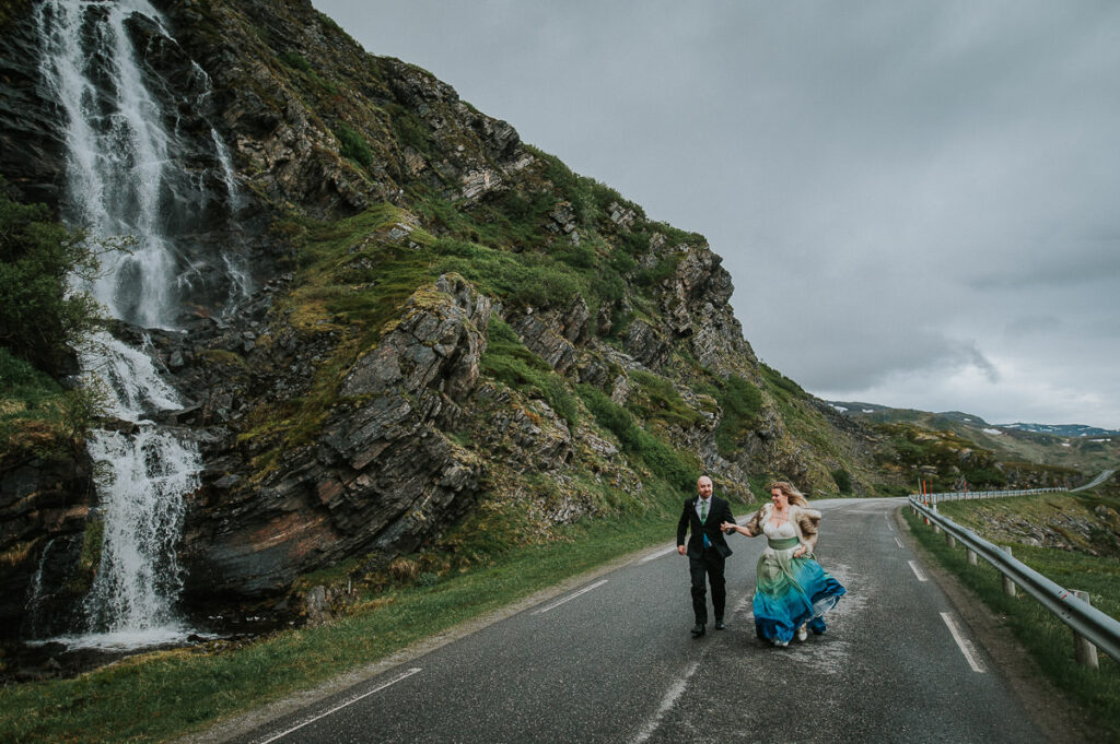 Bride and groom running in the middle of the road next to a waterfall in Sørøya island in Norway. Bride is wearing blue green wedding gown and has a viking inspired hair style