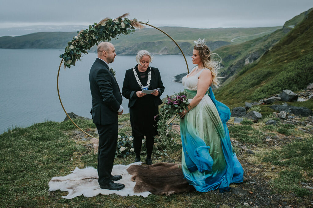 midnight sun elopement ceremony on Sørøya island in Norway. The couple is standing in front of a round wedding arch decorated with blue and green flowers. Bride is wearing a blue green wedding dress and has a viking inspired hair style