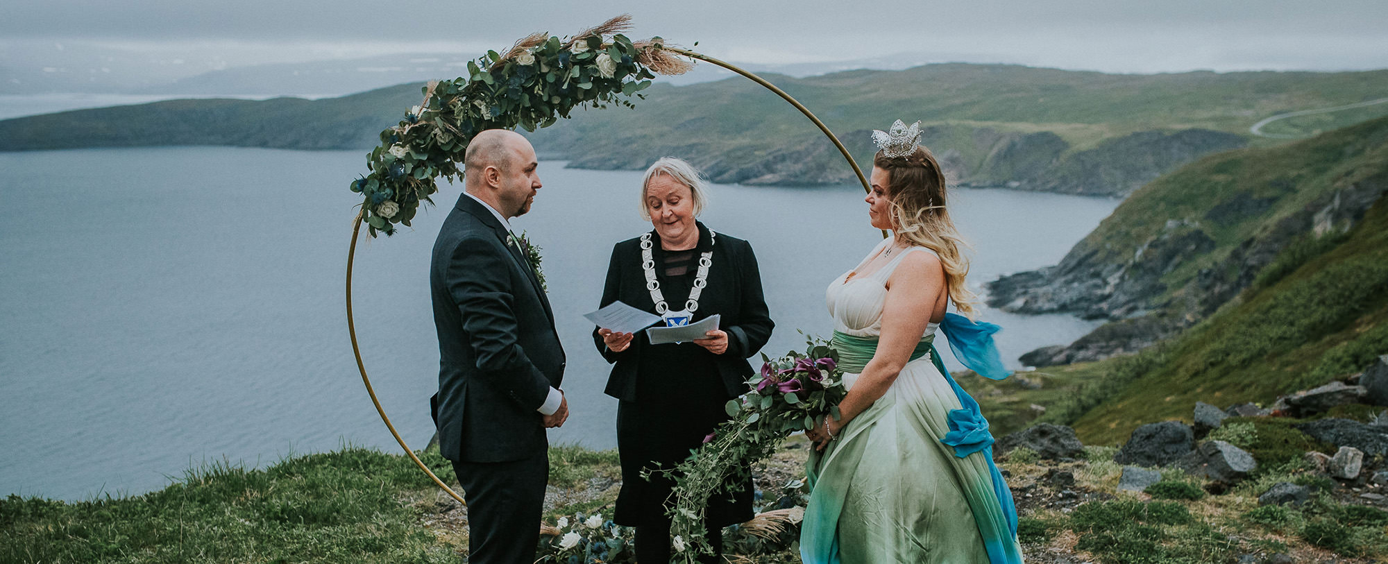 Gorgeous elopement ceremony on a distand island in Northern Norway. Bride is wearing a blue green dress and they are standing in front of a circular flower arch on their elopement day in Sørøya