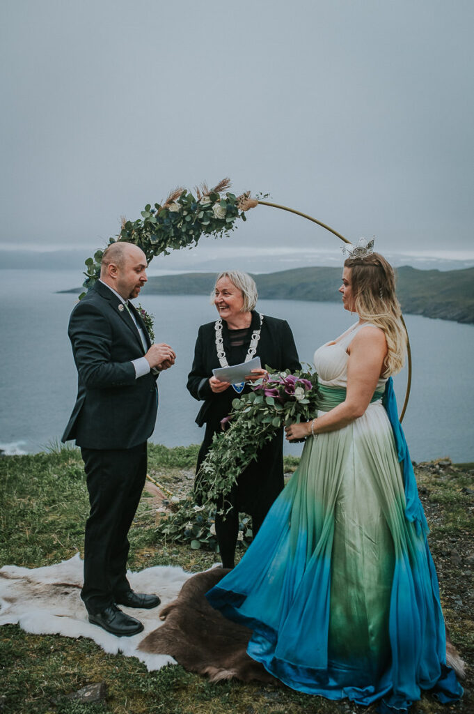 midnight sun elopement ceremony on Sørøya island in Norway. The couple is standing in front of a round wedding arch decorated with blue and green flowers. Bride is wearing a blue green wedding dress and has a viking inspired hair style