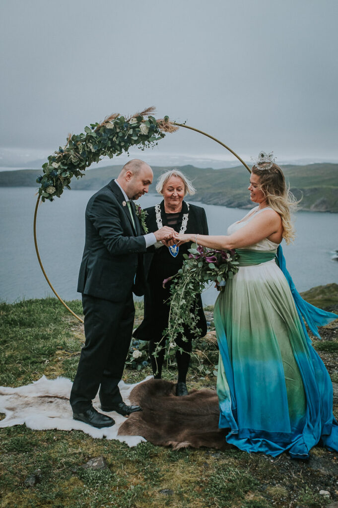 A bridal couple exchanging rings during their  elopement ceremony on Sørøya island in Norway. The couple is standing in front of a round wedding arch decorated with blue and green flowers. Bride is wearing a blue green wedding dress and has a viking inspired hair style