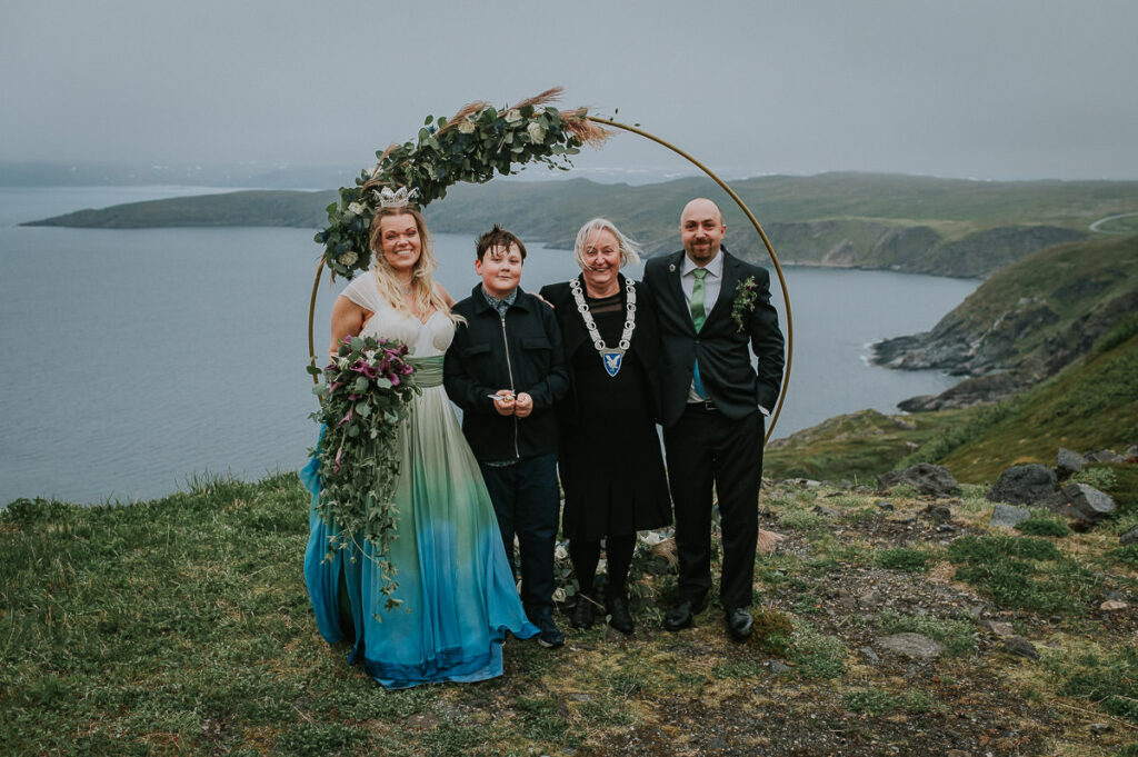 Midnight sun elopement ceremony on Sørøya island in Norway. The couple is standing in front of a round wedding arch decorated with blue and green flowers. Bride is wearing a blue green wedding dress and has a viking inspired hair style