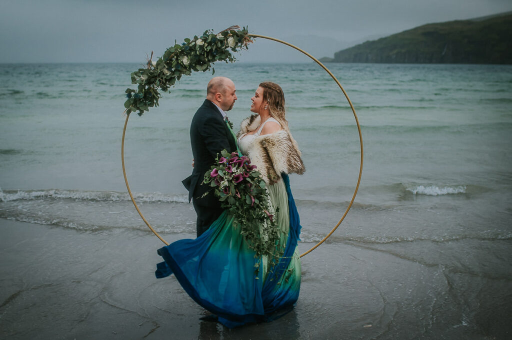 Bride and groom hugging in front of a round floral arch decorated  with blue and green flowers in the sea on a foggy night . They are dipping toes in the cold water and enjoying their midnight sun elopement in Sørøya island in Norway