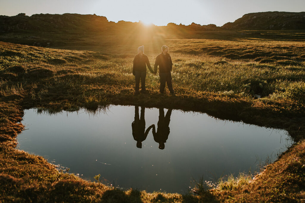 A couple holding hands and  walking in front of a lake and they are mirrored in the water while midnignt sun is shining behind them on Sørøya island in Norway