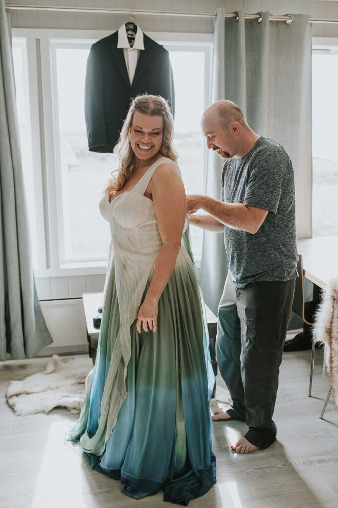 Groom is helping bride to get the dress on. The dress is ivory colored with blue and green details