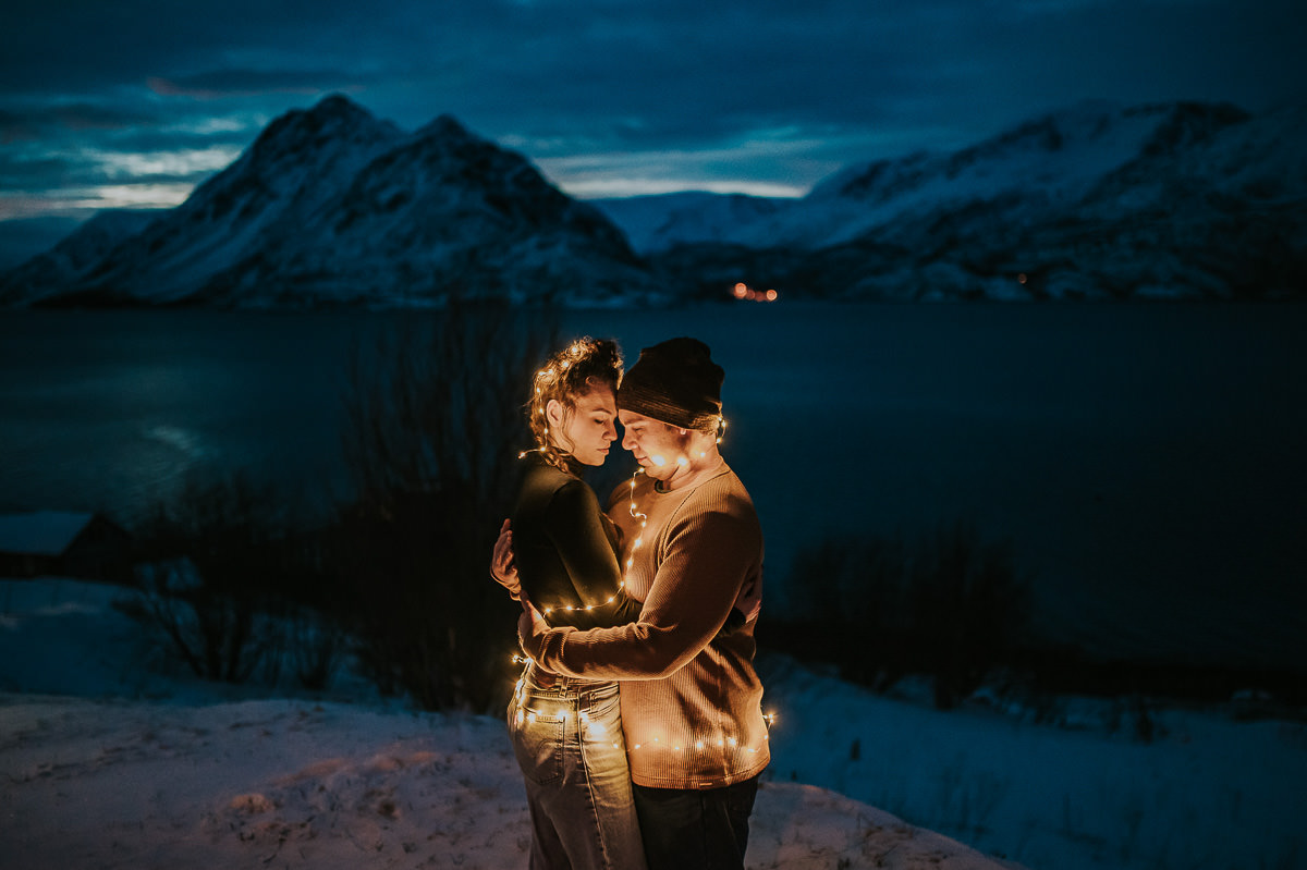 A couple with fairy lights on them posing in front of a winter mountain landscape in Alta Norway
