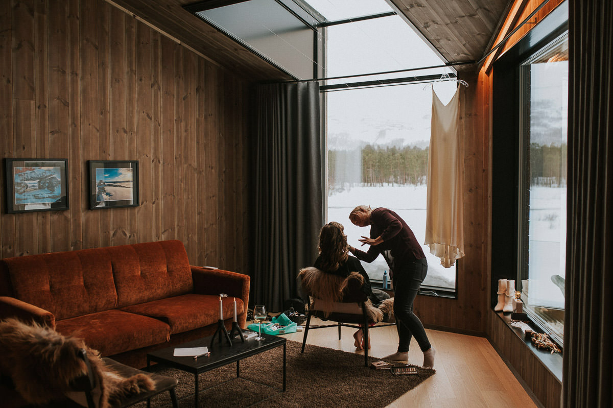 A bride getting ready for her winter elopement day in Alta Norway. She is getting her makeup done in front of a big window with a snowy landscape outside