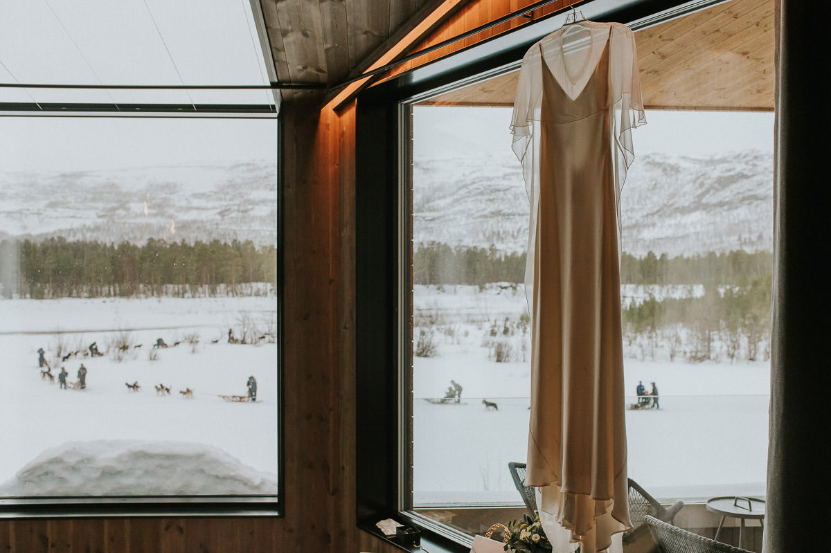 Beautiful wedding gown with a silk cape hanging in front of a big window. Outside there is a winter storm and there are several dogsledding teams outside mushing through the fresh snow