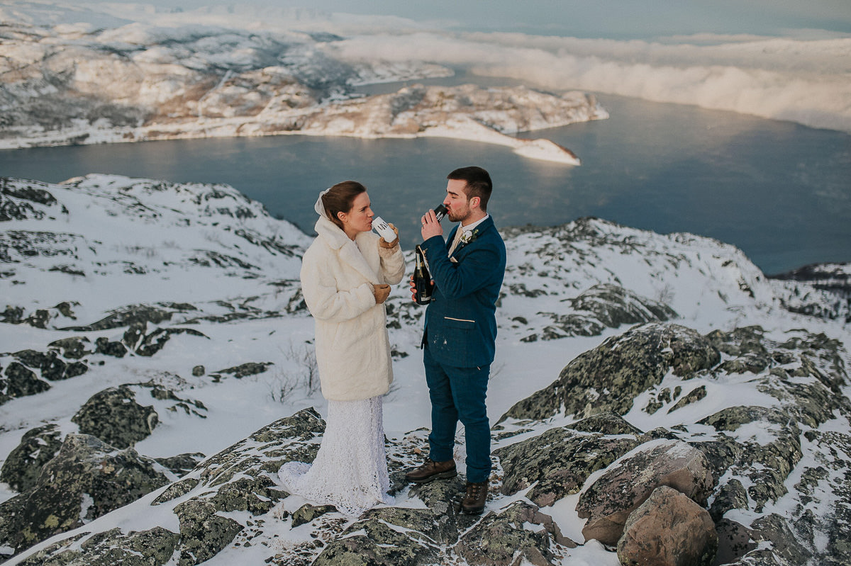 Bride and groom on a snow-capped mountaintop drinking champagne to celebrate their elopement ceremony in Alta Norway