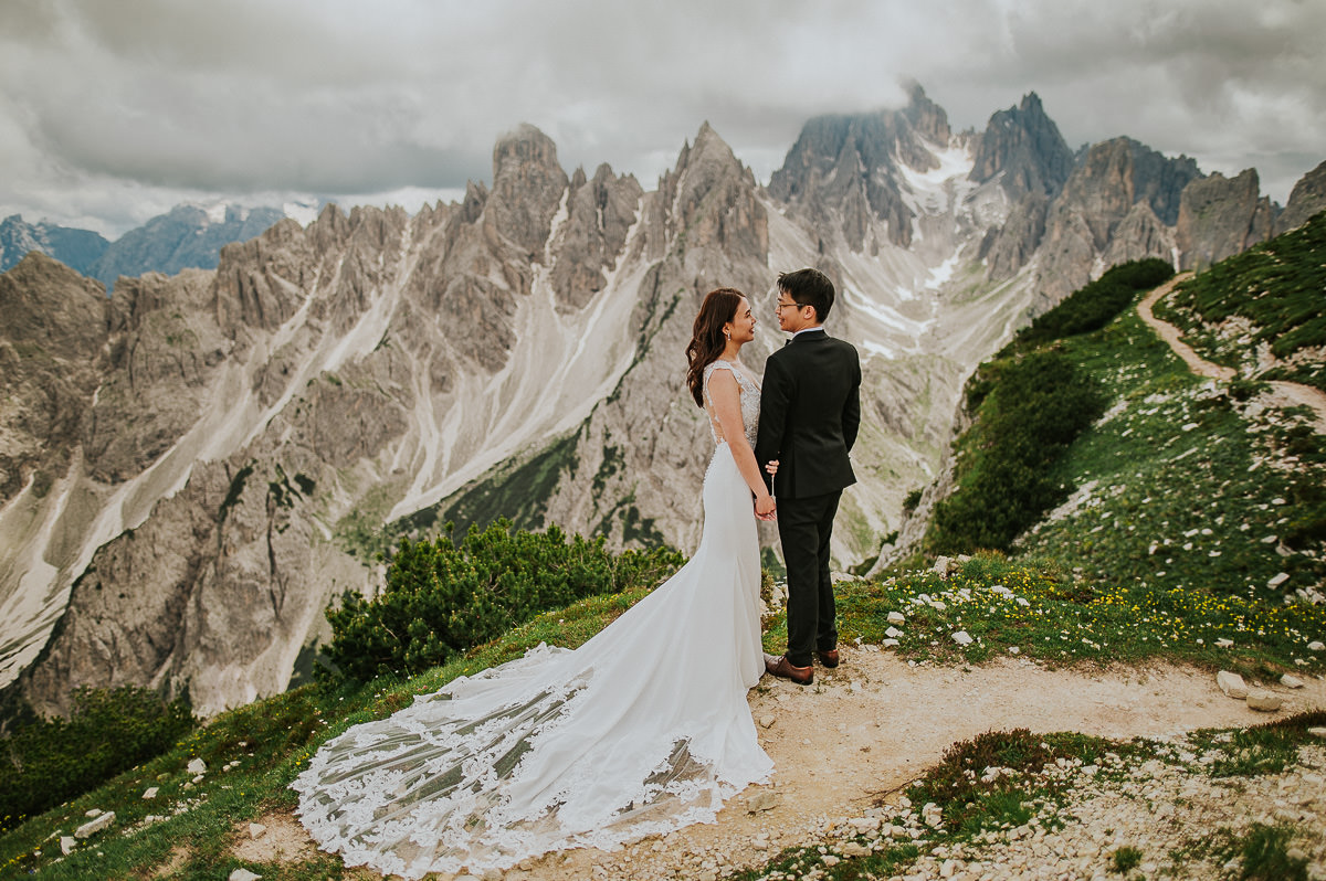 Bride and groom walking in the mountains Tre Cime di Lavaredo in Italian Dolomites on a sunny summer day. The bride is wearing a lace wedding gown with a long train 