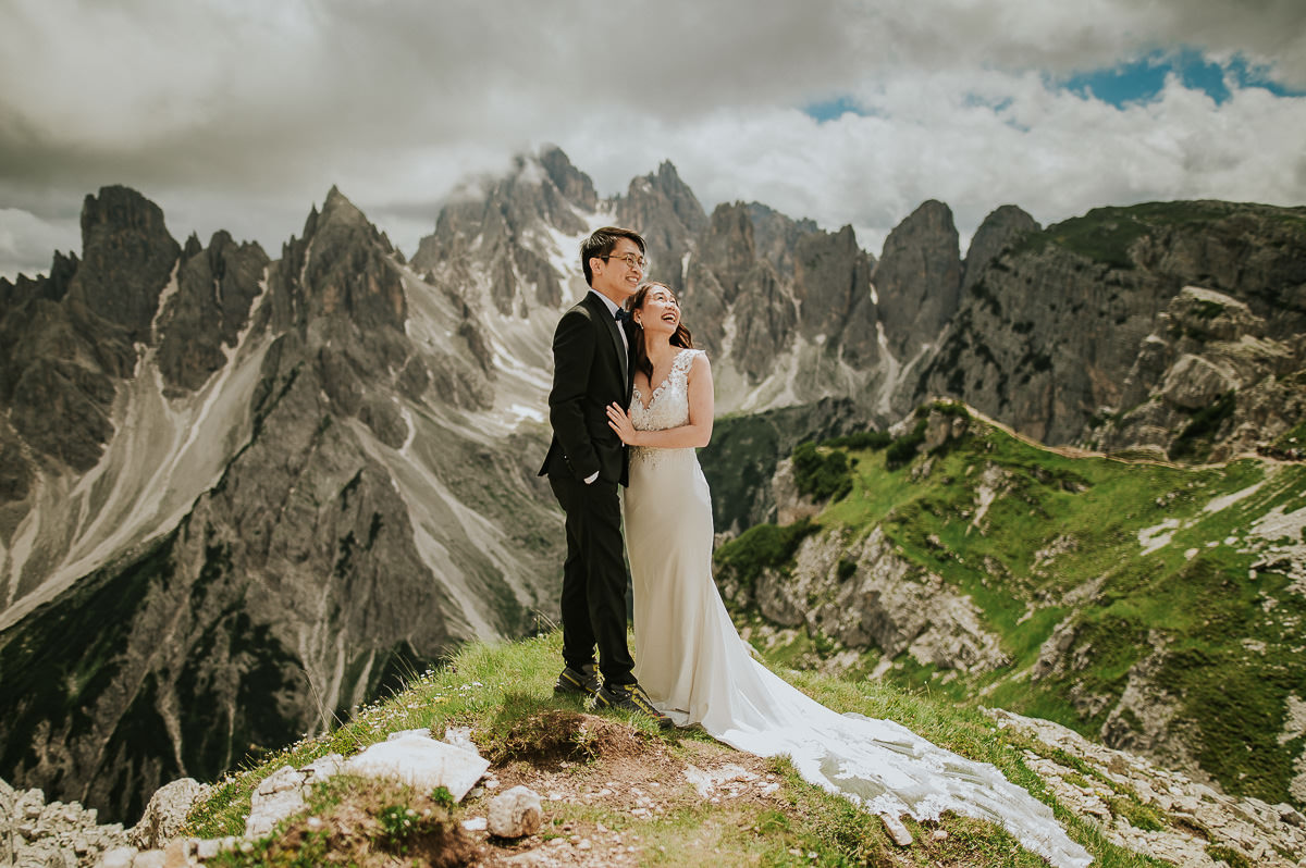 Bride and groom in the mountains Tre Cime di Lavaredo in Italian Dolomites on a sunny summer day. The bride is wearing a lace wedding gown with a long train 