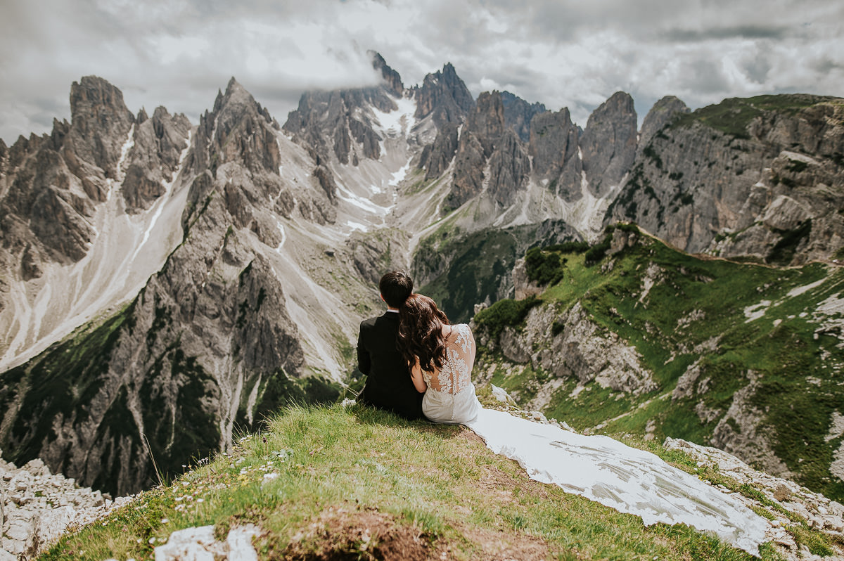 Bride and groom sitting on a cliff in the mountains Tre Cime di Lavaredo in Italian Dolomites on a sunny summer day. The bride is wearing a lace wedding gown with a long train  