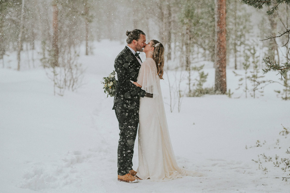 Gorgeous bride and groom kissing in a forest under a heavy snowfall on the day of their winter elopement in Alta Norway