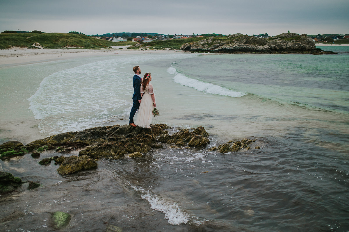 Utendørs sommer bryllup i Haugesund - brudeparet tar bilder på en vakker strand med turkis vann og bølger i bakgrunn