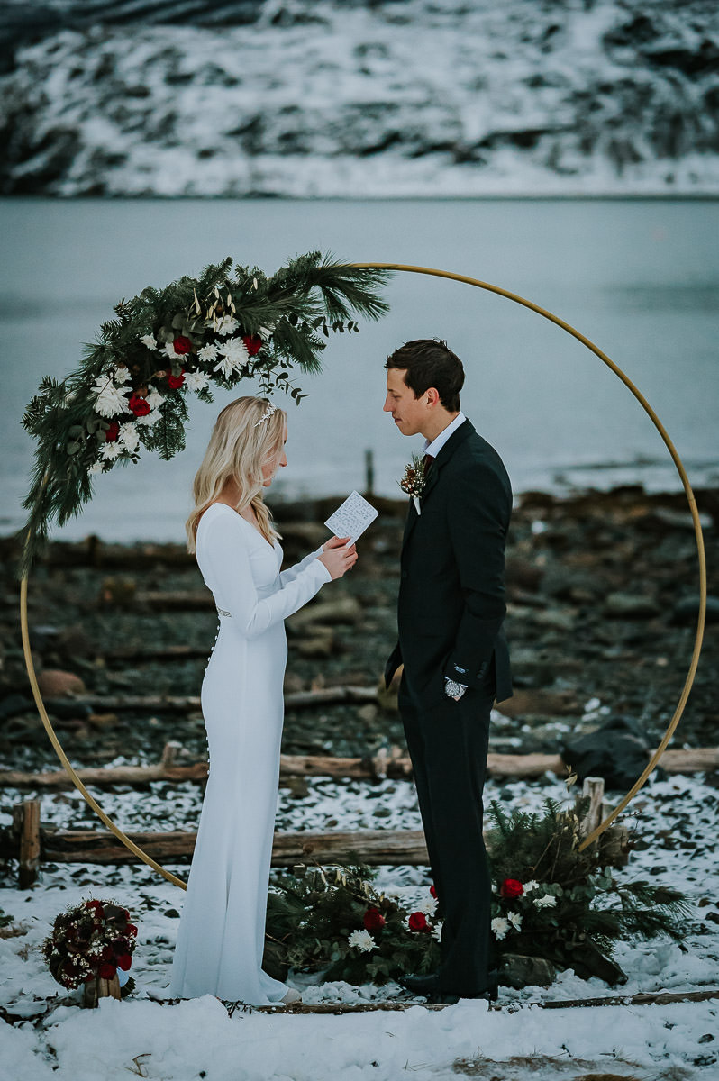 Utendørs bryllupsseremoni foran havet  i Alta på en nydelig vinterdag hvor brudeparet leser løftene sine for hverandre. Bak dem er det en rund flott blomsterbrue dekorert med friske blomster i hvit og rød farge