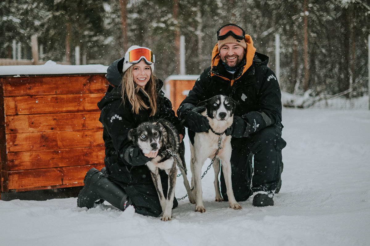 Adventure couple in winter outfits cuddling with dogs on a husky farm under a heavy snowfall