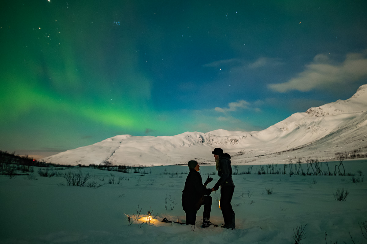 Surprise proposal under the northern lights in Tromsø Norway with aurora borealis dancing in the sky and snow-capped mountain landscape in the background - a guy is standing on his knee before his girlfriend