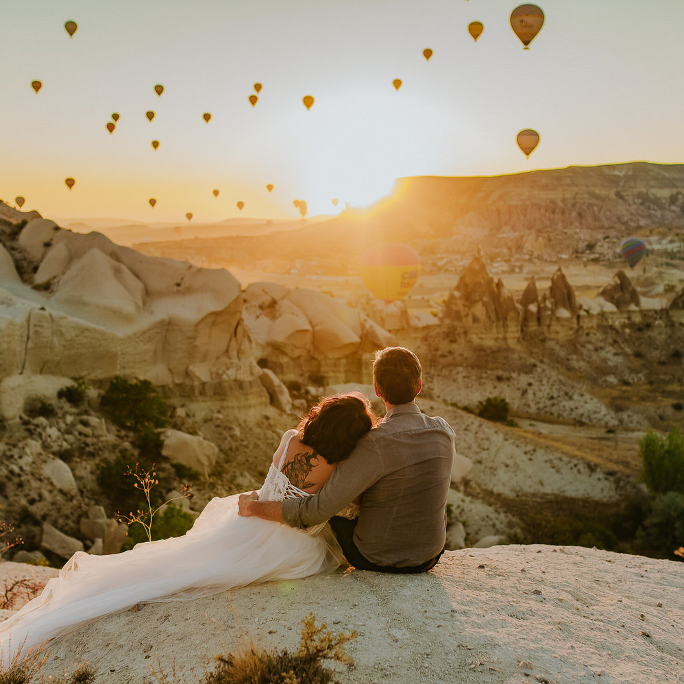 Beautiful couple in wedding attire enjoying sunrise and hot air balloons rising above mountains in Cappadocia Turkey