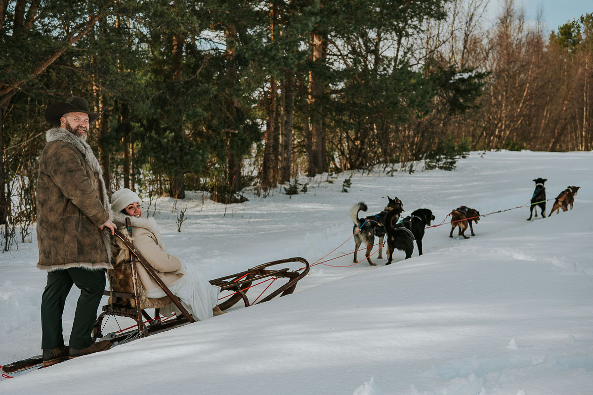 Bride and groom in their wedding attire dog sledding in the woods of Alta Norway on the day of their destination adventure wedding