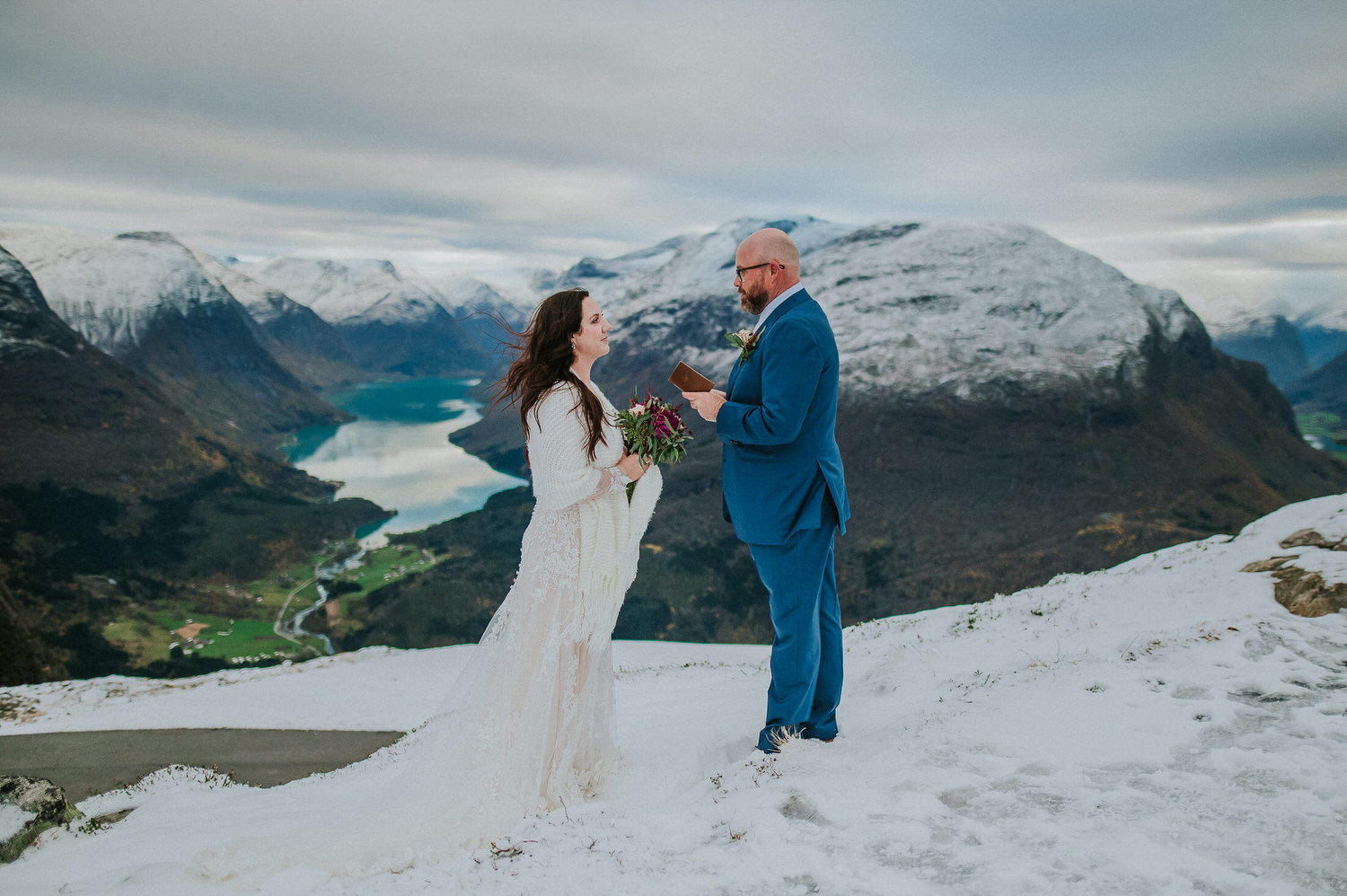Beautiful couple in wedding attire reading their vows to each other on a mountaintop among stunning winter landscapes of Loen in Norway