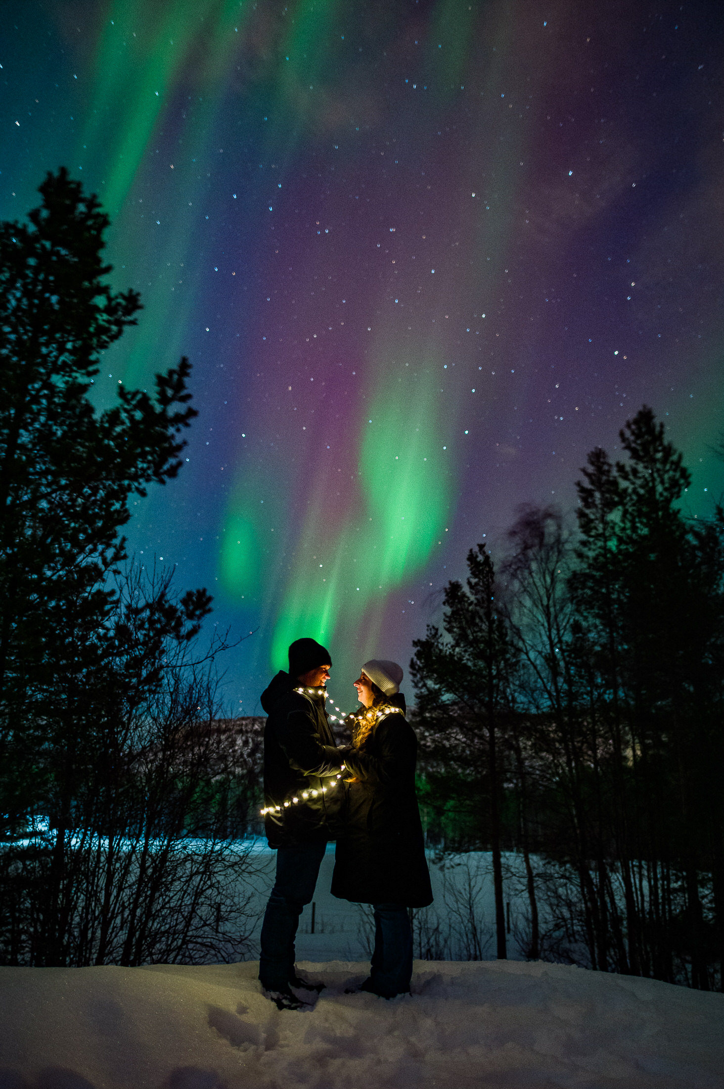 Sweet couple with fairy lights and northern lights in a beautiful winter forest in Alta Norway on the day of their engagement photo session