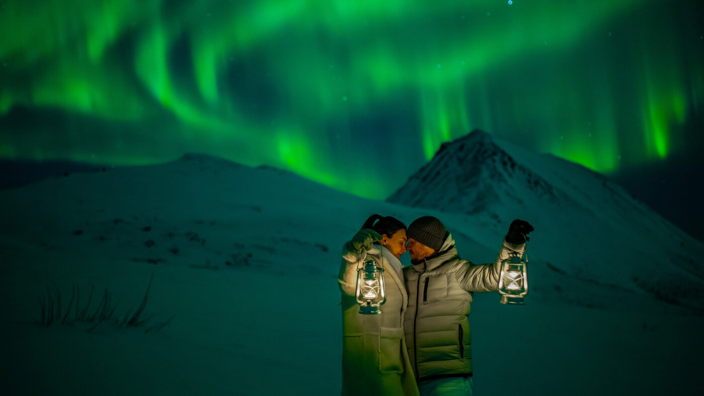 Beautiful and romantic engagement photo session under the northern lights in Tromsø Norway - the couple is cuddling together while watching aurora borealis dancing above snowy mountains