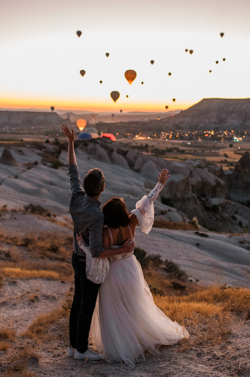 Bride and groom at dawn in Cappadocia waving to hot air balloons drifting in the sky
