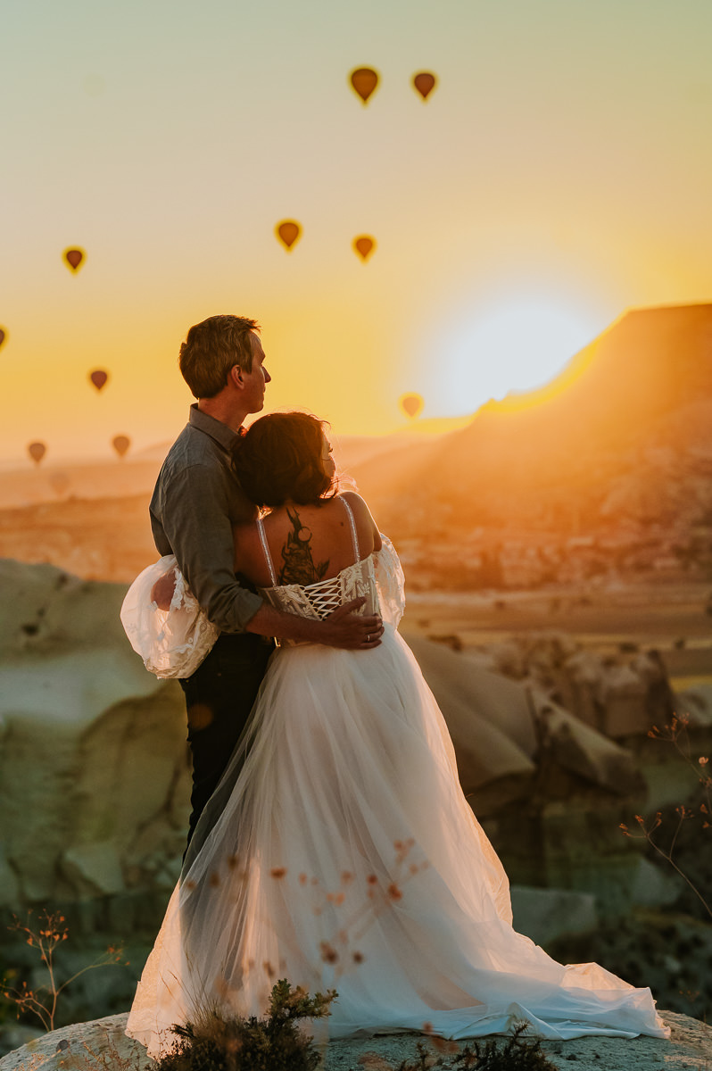 Bride and groom watching a stunning sunrise and hot air balloons drifting in the sky in Cappadocia on the day of their elopement