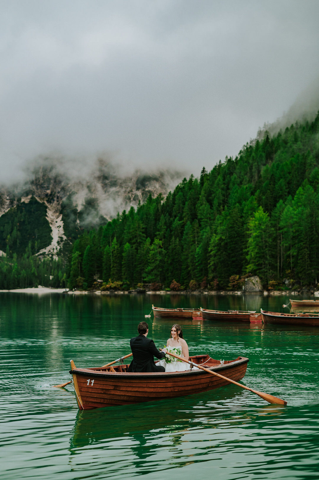 Bride and groom rowing an old wooden boat at Lago di Braies in the Italian Dolomites on the day of their adventure elopement - captured by elopement photographer TS Foto Design