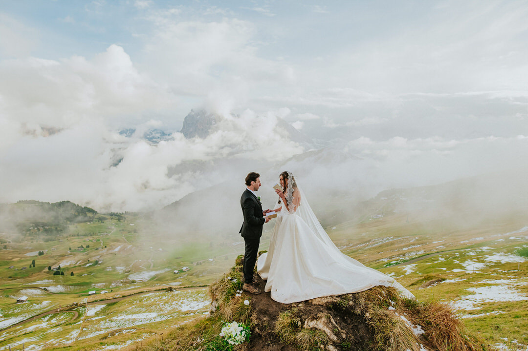 Seceda elopement in the Italian Dolomites - bride and groom reading their vows to each other on the mountaintop surrounded by clouds 