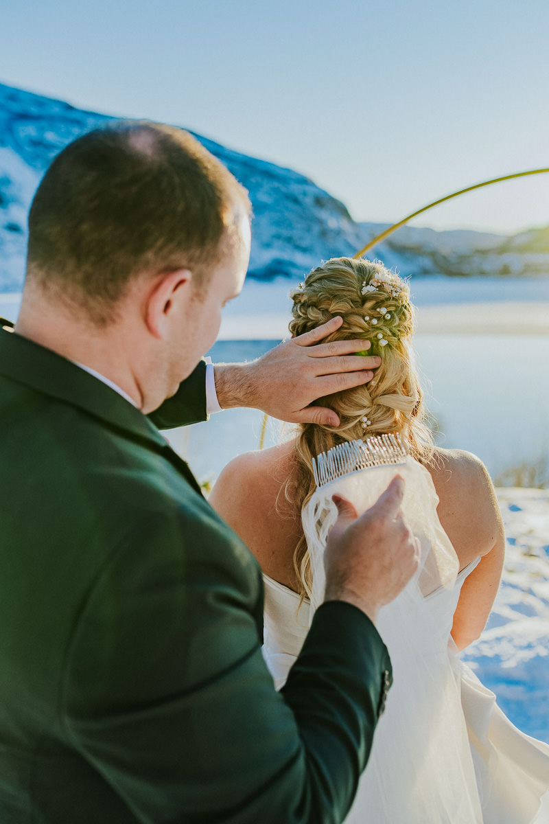 Groom is helping the bride to put on a veil before their winter elopement ceremony in Arctic Norway
