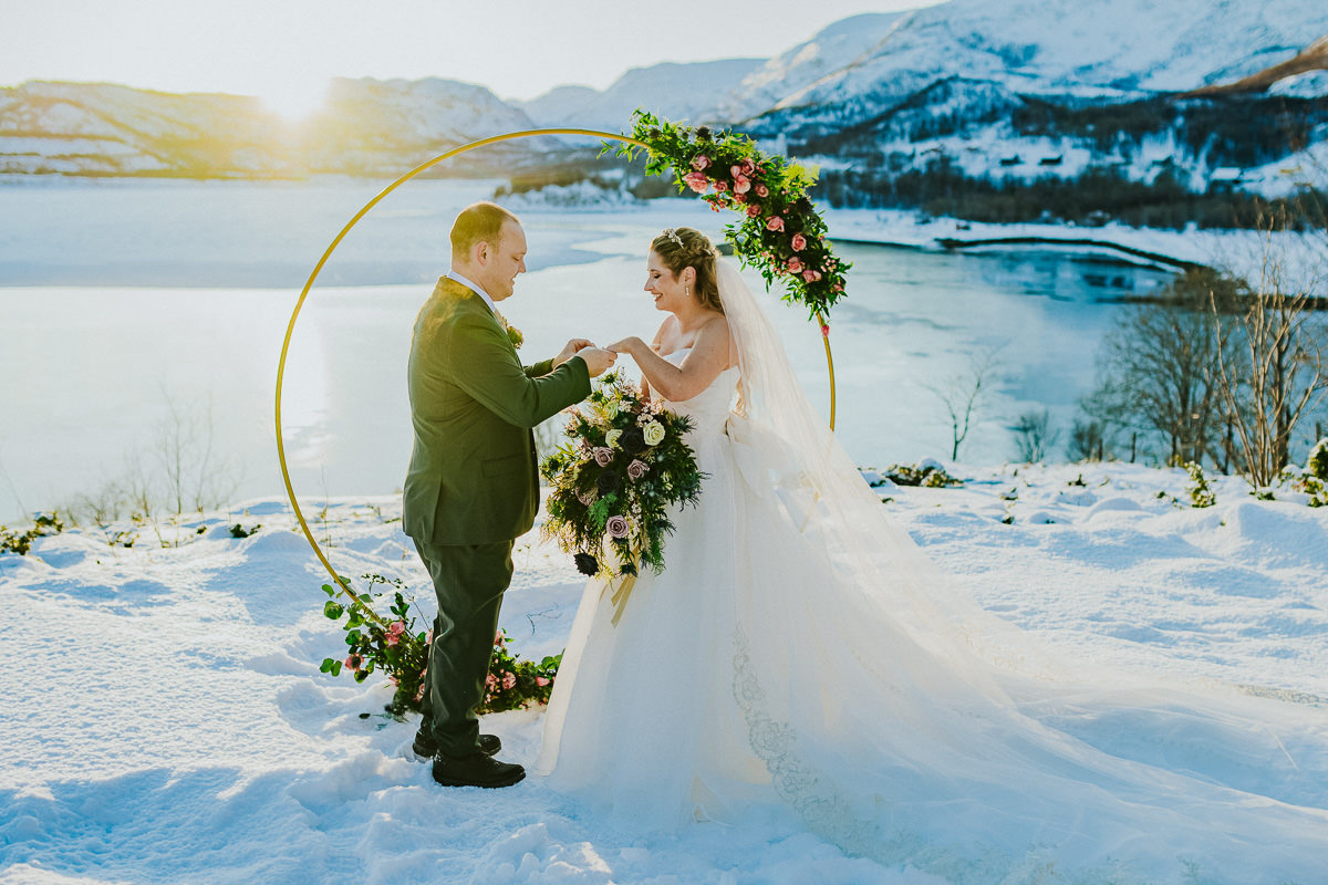 Bride and grom exchanging their rings on the day of their winter arctic elopement in Alta Norway. THere is a fjord and snowy mountain landscapes in the backgroun and the golden light of the sunset is shining through