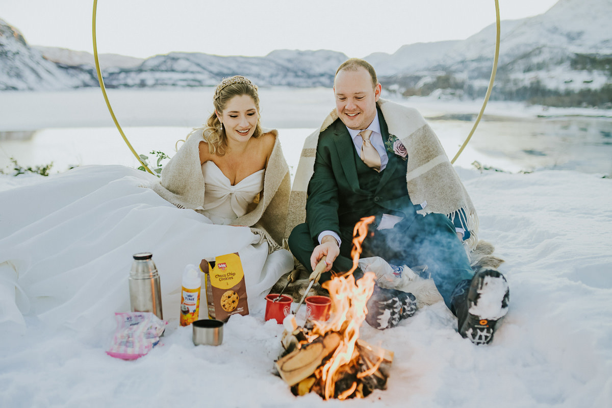 Bride and groom sitting by a campfire on the day of their winter elopement in Alta Norway  and making s´mores