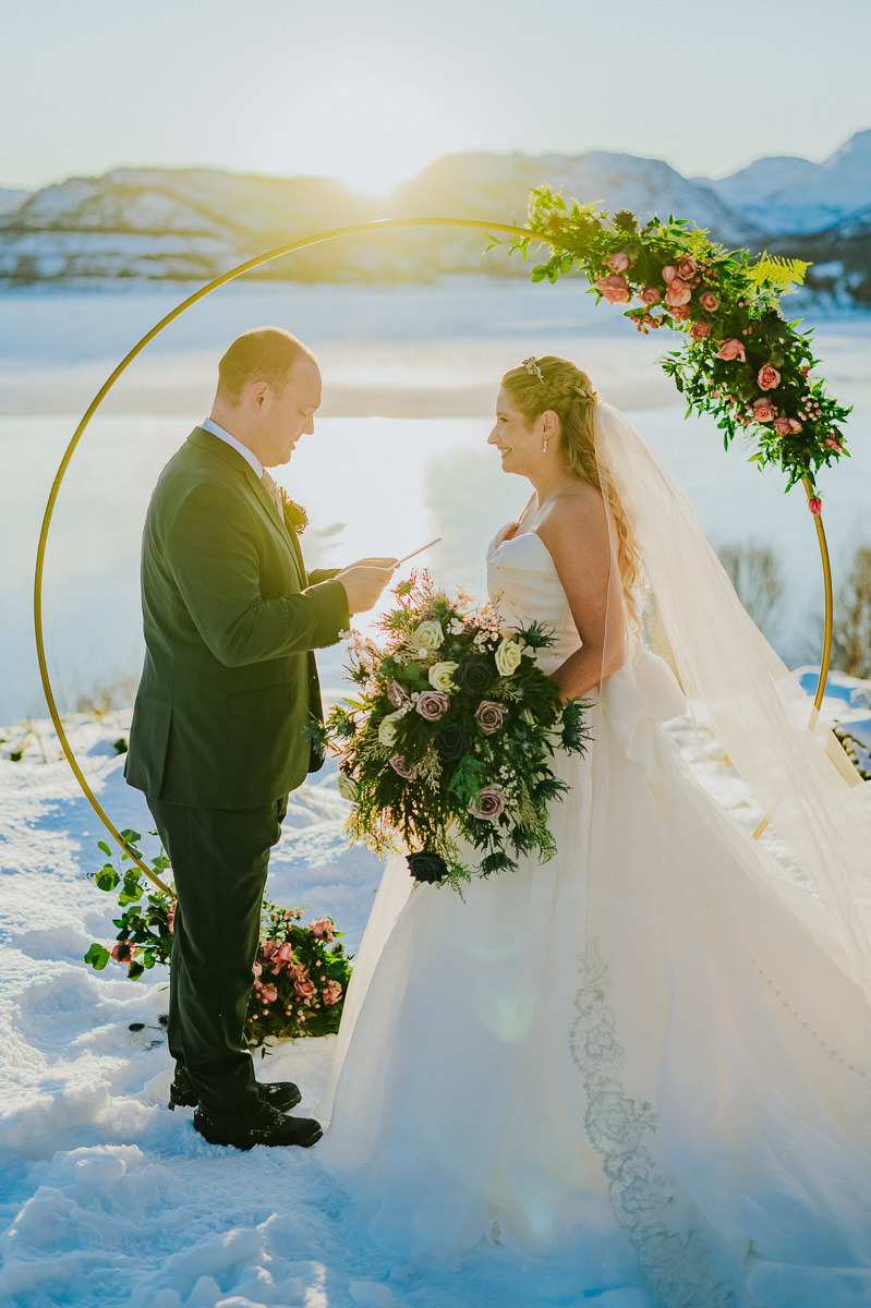Bride and grom reading their vows to each other on the day of their winter arctic elopement in Alta Norway. THere is a fjord and snowy mountain landscapes in the backgroun and the golden light of the sunset is shining through
