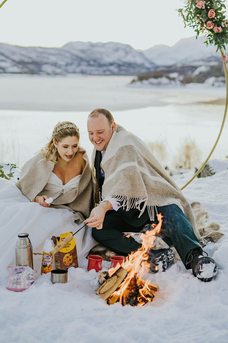 Bride and groom sitting by a campfire on the day of their winter elopement in Alta Norway  and making s´mores
