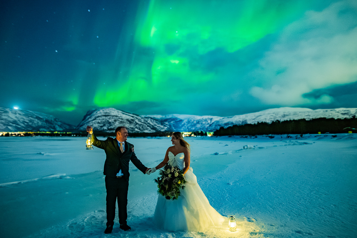 Spectacular portrait of a bride and groom in front of snowy mountains and the sea with dancing green northern lights above them - aurora borealis wedding in Alta Norway captured by adventure elopement photographer TS Foto Design