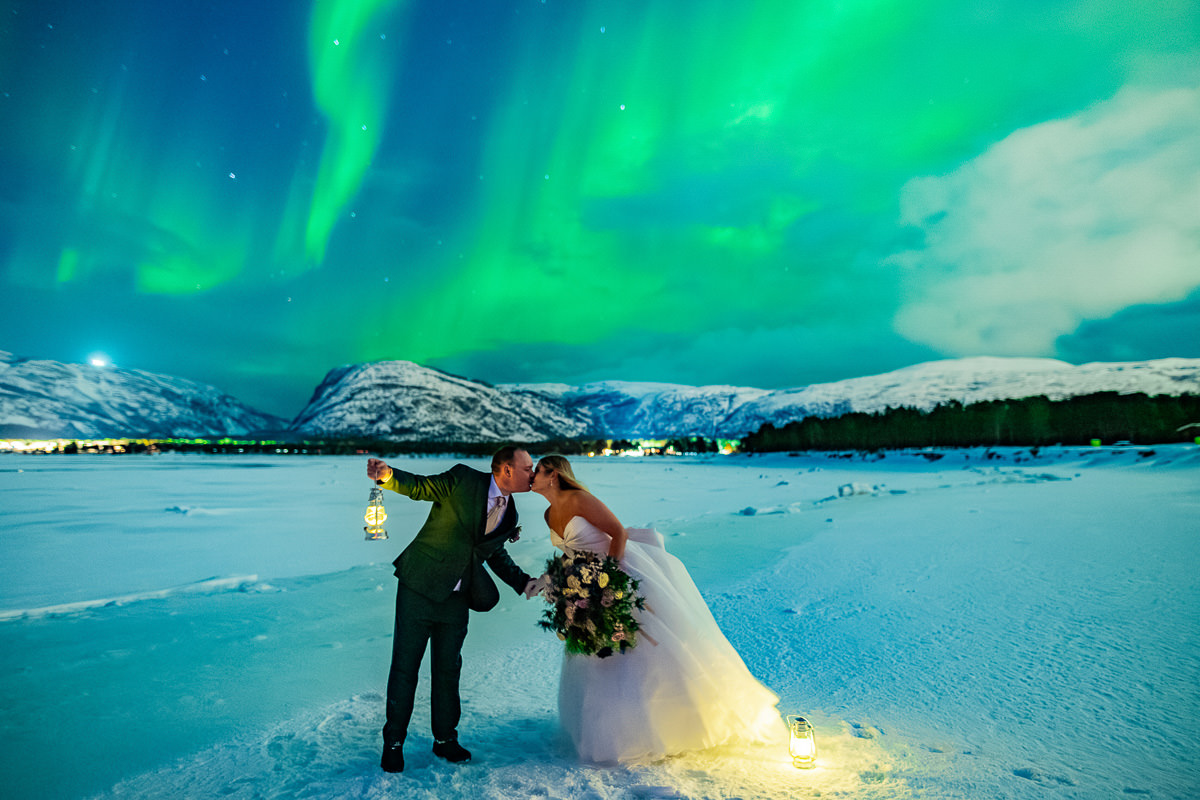 Epic portrait of a bride and groom in front of snowy mountains and the sea with dancing green northern lights above them - aurora borealis wedding in Alta Norway captured by adventure elopement photographer TS Foto Design