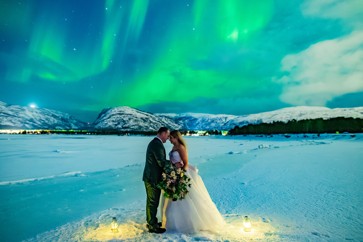 Spectacular portrait of a bride and groom in front of snowy mountains and the sea with dancing green northern lights above them - aurora borealis wedding in Alta Norway captured by adventure elopement photographer TS Foto Design