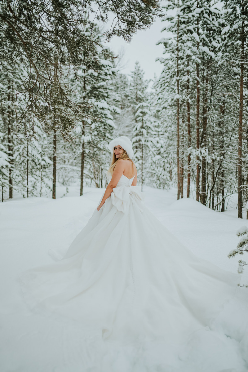 Gorgeous bride in a big wedding dress with a bow wearing white winter hat in a snow covered forest in Alta Norway