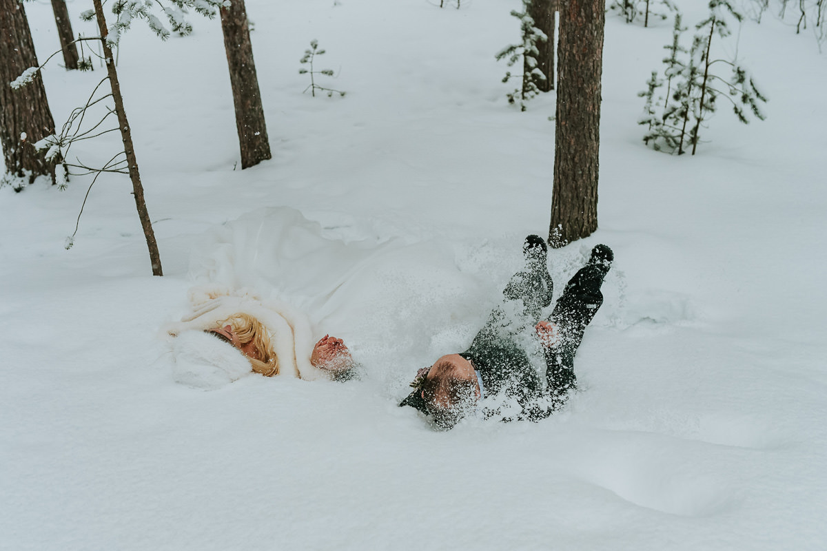Bride and groom in their wedding attire falling in the snow in a winter forest in Alta Norway on the day of their arctic wedding