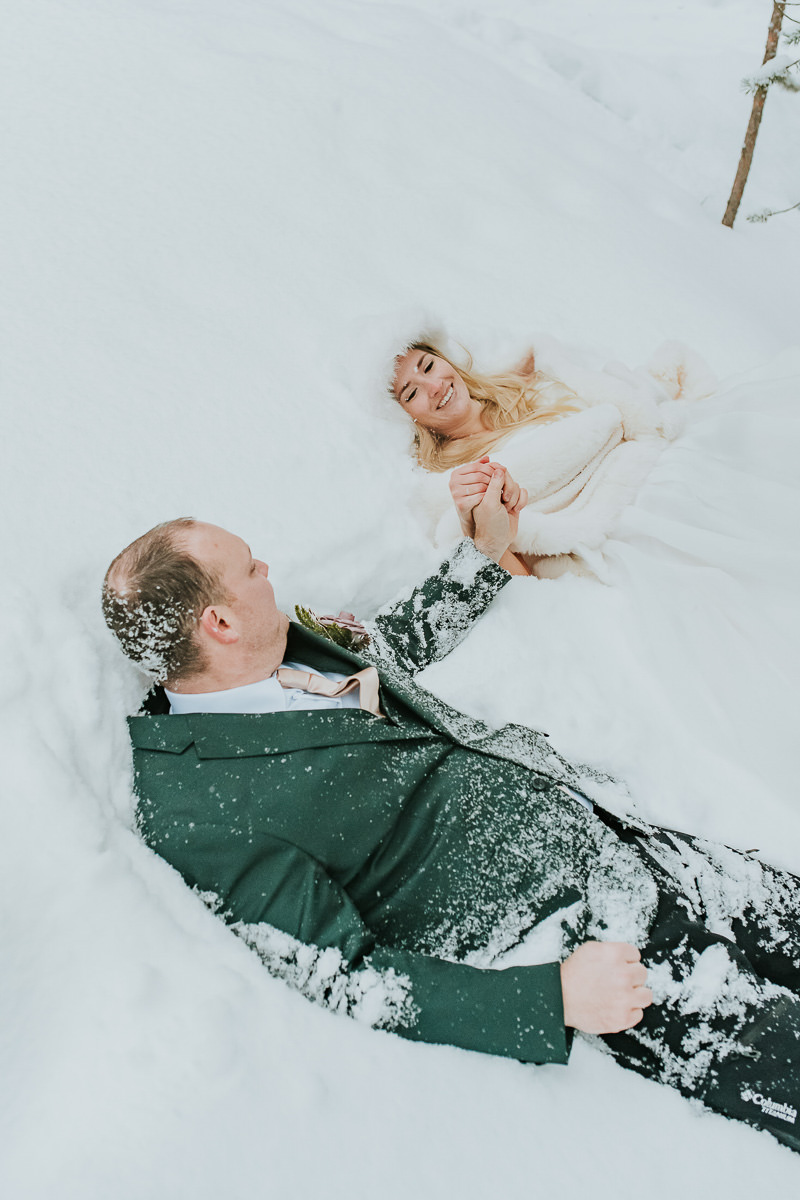 Bride and groom in their wedding attire falling in the snow in a winter forest in Alta Norway on the day of their arctic wedding