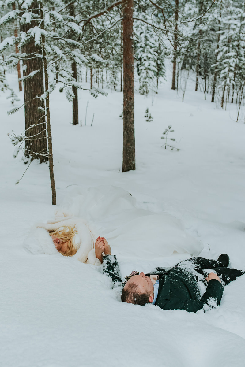 Bride and groom in their wedding attire falling in the snow in a winter forest in Alta Norway on the day of their arctic wedding