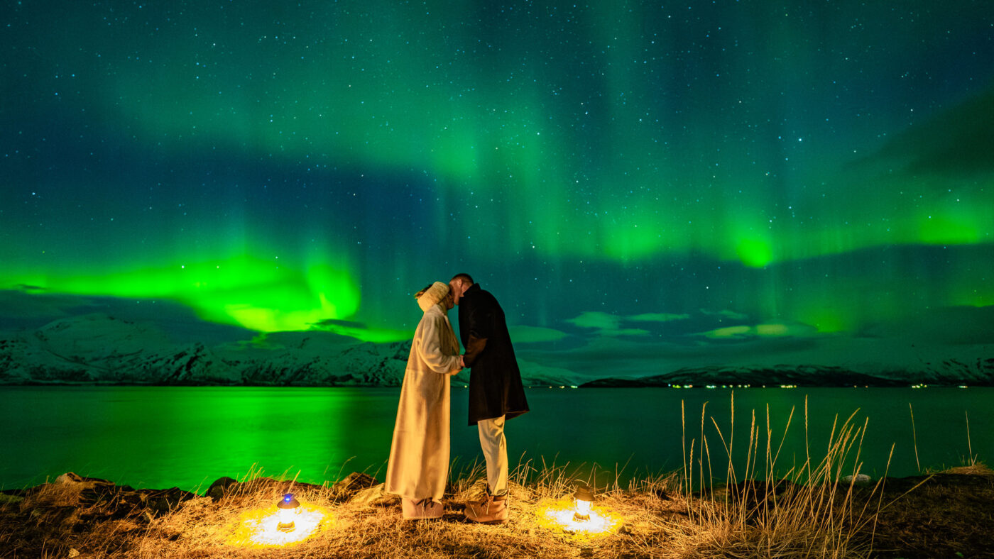 Stunning portrait of a couple kissing under the northern lights in Tromsø Norway. There are white snowy mountains in the background and the aurora is nicely reflected in the fjord behind the couple