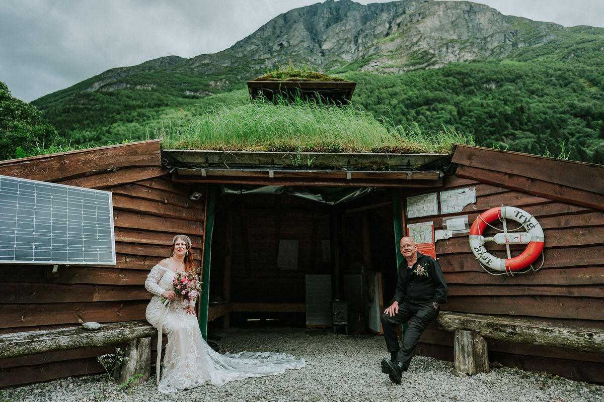 Bride and groom sitting outside on wooden benches in front of a grassy-roof cabin in Norway